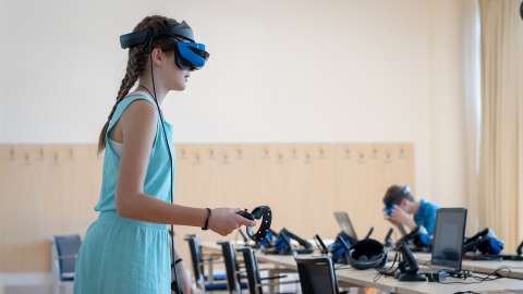 Schülerinnen und Schüler, die vor dem Schreibtisch stehen, ein VR-Headset tragen und den Controller in einer Hand halten, während sie an einer VR-basierten Lektion teilnehmen