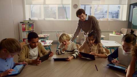 Sechs junge Schüler mit Tablets sitzen an einem Tisch im Klassenzimmer und ein Lehrer blickt einem Schüler bei der Arbeit über die Schulter.