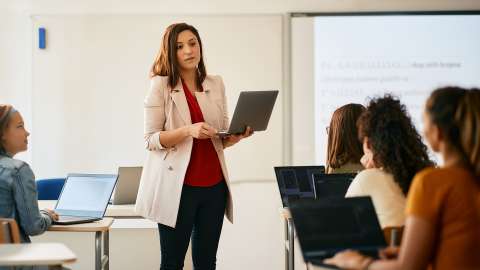 Lehrer, der einen offenen Laptop hält, während er vor einer Klasse mit Schülern steht, die alle einen Laptop bzw. ein Chromebook vor sich haben