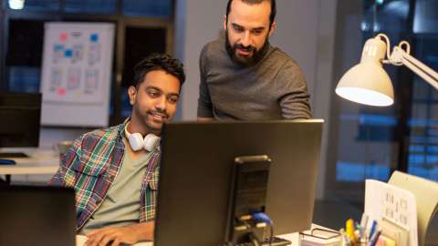 Zwei Kollegen in einem modernen, offenen Arbeitsbereich überprüfen gemeinsam Informationen, die auf einem Desktop-Monitor angezeigt werden. Eine Person sitzt und lächelt, während sie auf einer Tastatur tippt, während die andere steht und auf etwas auf dem Bildschirm zeigt